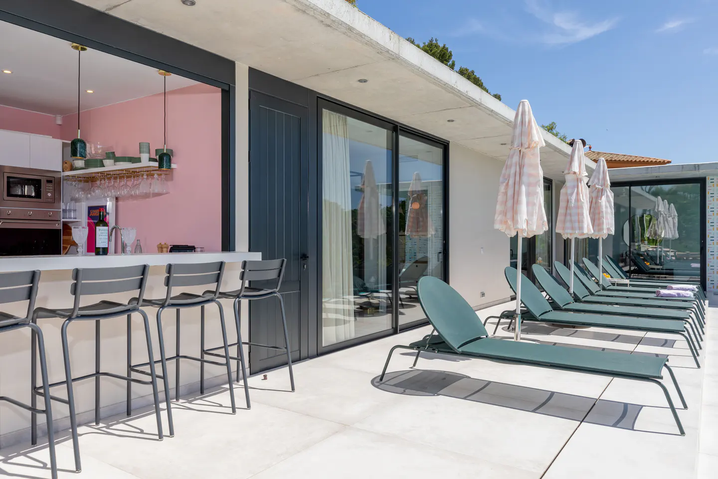 Outdoor bar and pool area with lounge chairs. Bar stools line a white counter with a pink wall behind. Lounge chairs sit under umbrellas.
