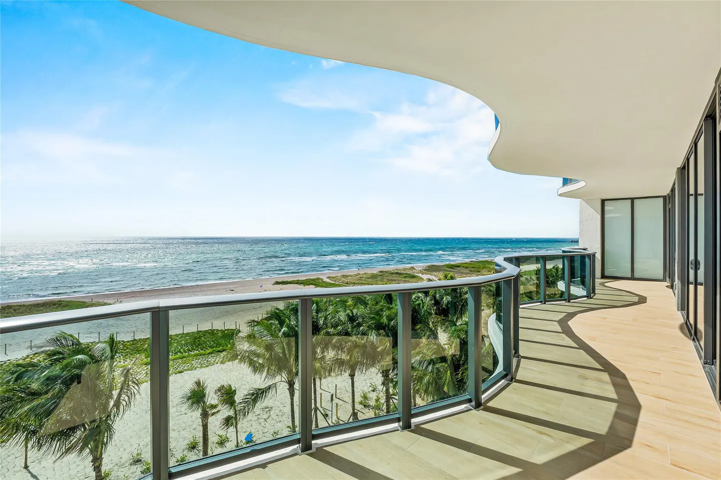 Ocean view from a curved balcony with glass railings. Palm trees and beach visible. Blue sky.