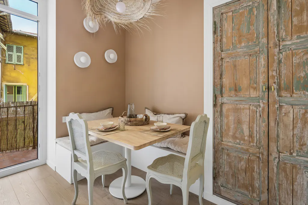A cozy dining nook with a square table set for two, surrounded by a built-in bench, chairs, and rustic wooden doors. Balcony view.