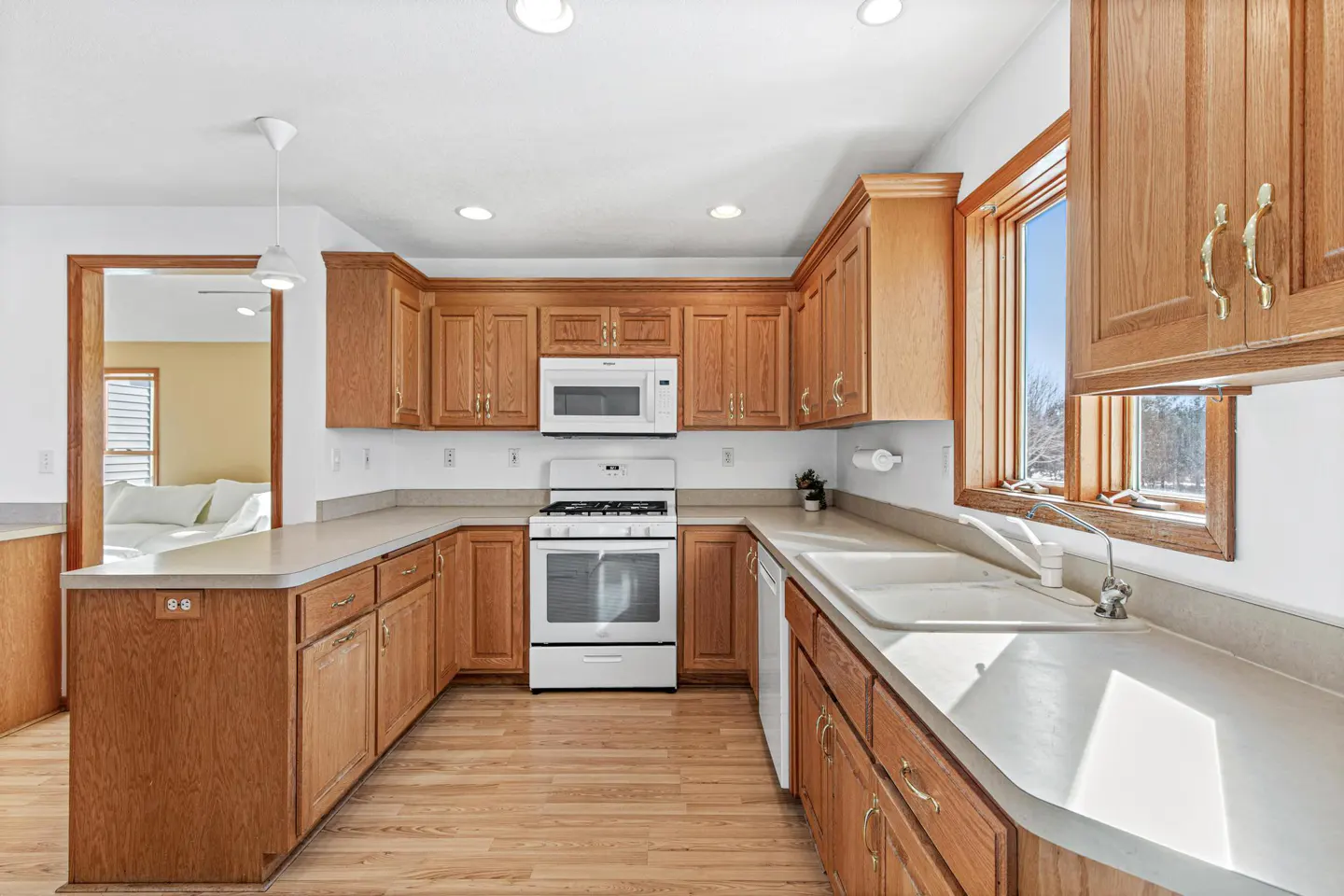 A bright kitchen with wood cabinets, white appliances, and light wood floors. A doorway leads to a room with a white sofa.