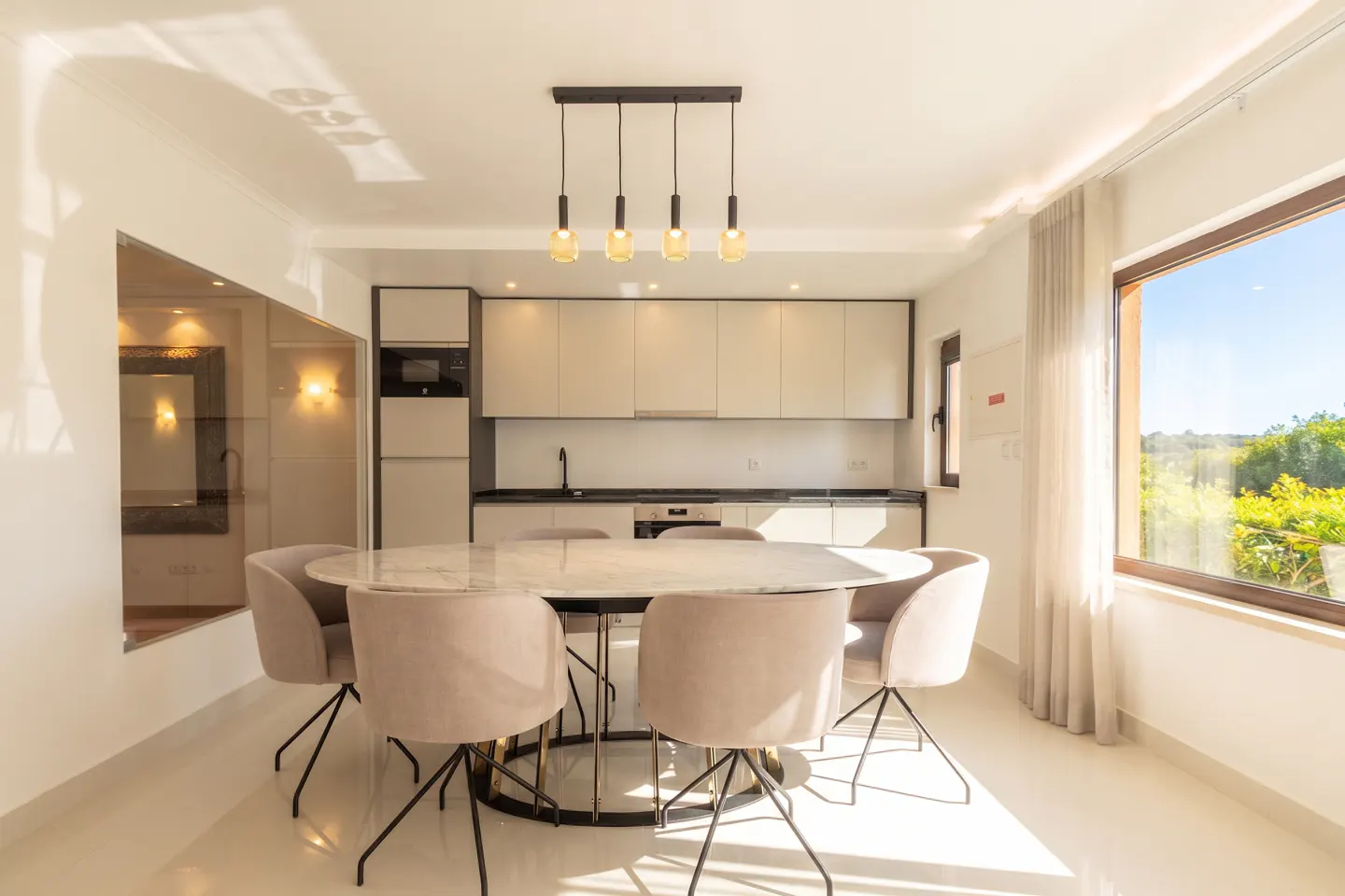 Bright, modern kitchen with an oval marble table surrounded by six beige chairs under a four-bulb pendant light. A large window shows a green landscape.