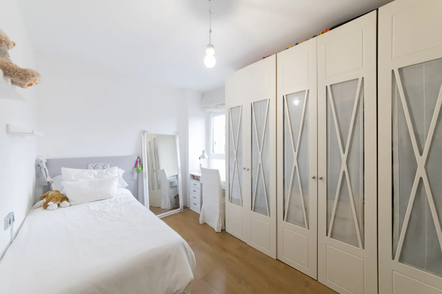 Bright bedroom with a white bed, desk, and large wardrobe with glass-paneled doors. A full-length mirror stands near the bed.