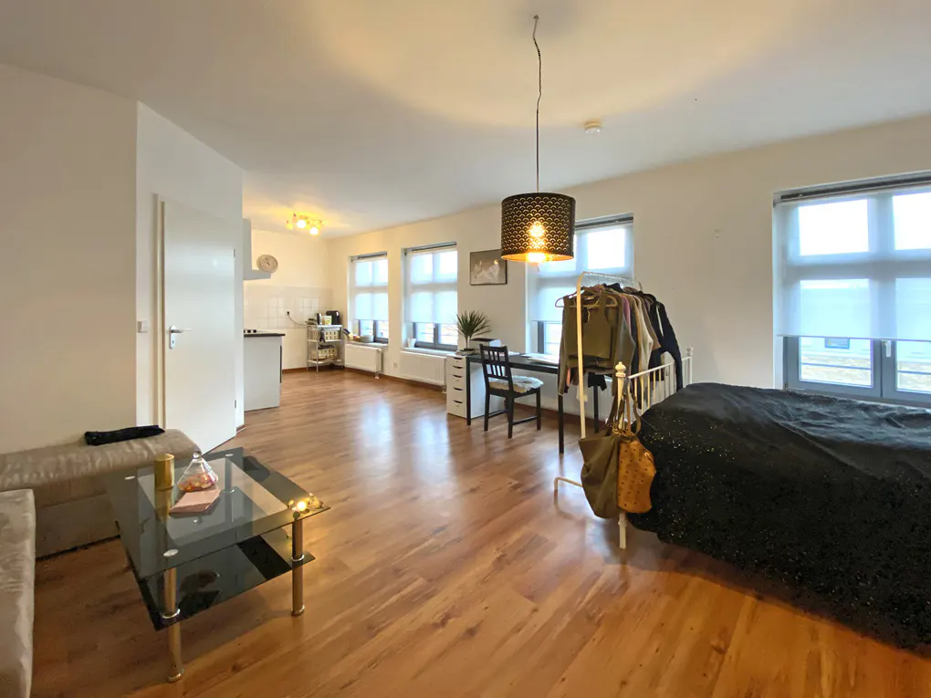 Bright studio apartment with wood floors, white walls, and large windows. A black bedspread is visible, along with a glass coffee table and kitchen area.