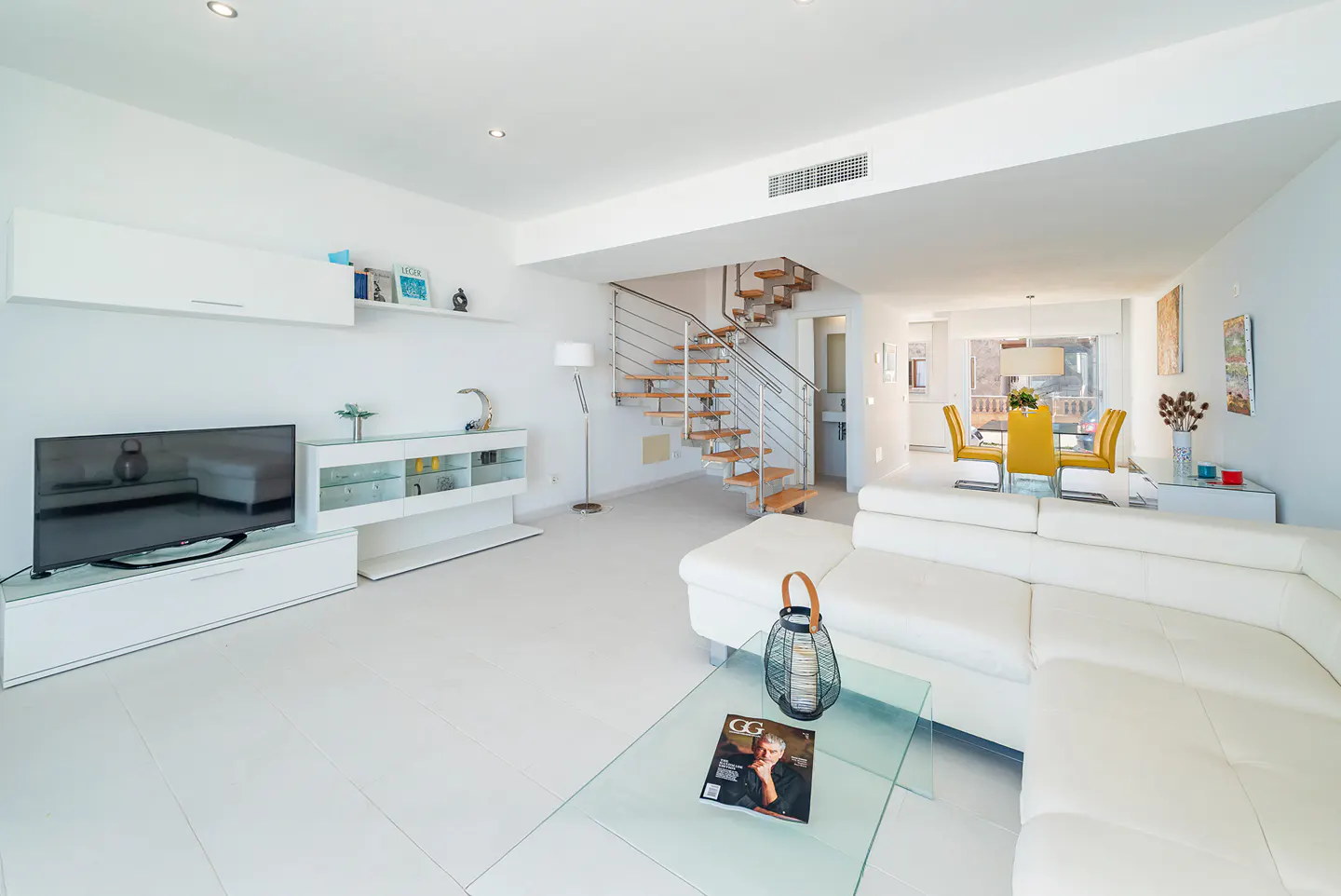 Bright, modern living room with white walls, floors, and furniture. A glass coffee table sits in front of a white sectional sofa. Stairs lead to the upper level.