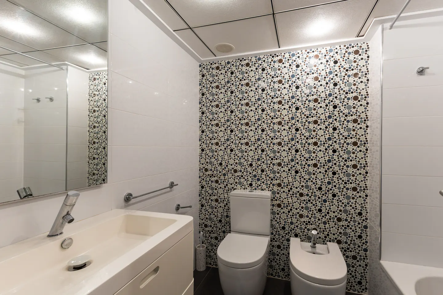 Bright bathroom with white tiles, a mosaic accent wall, toilet, bidet, and a modern sink with a large mirror.