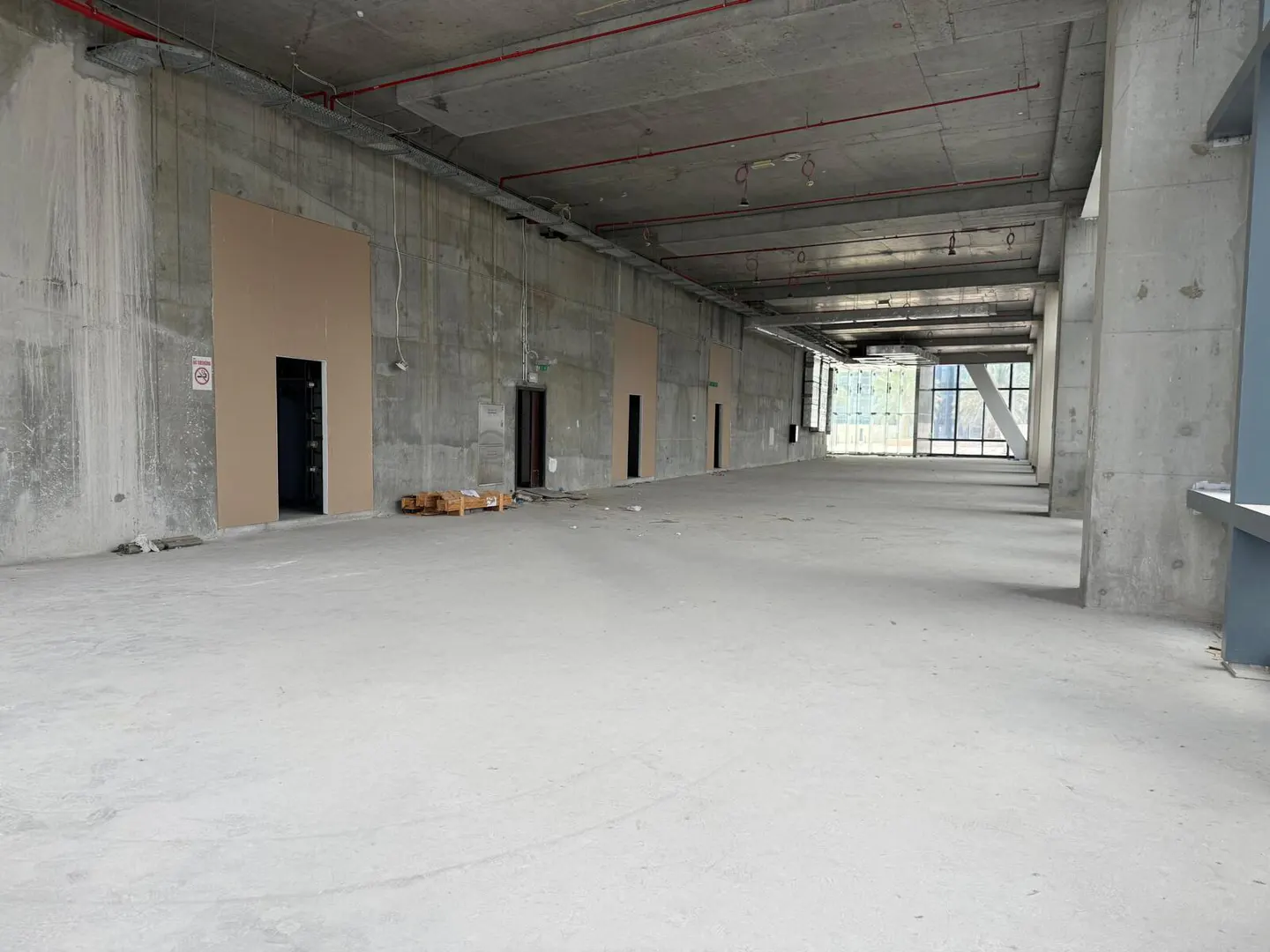 Unfinished commercial space with concrete walls, floor, and ceiling. Doors line the left wall, and windows are at the far end.