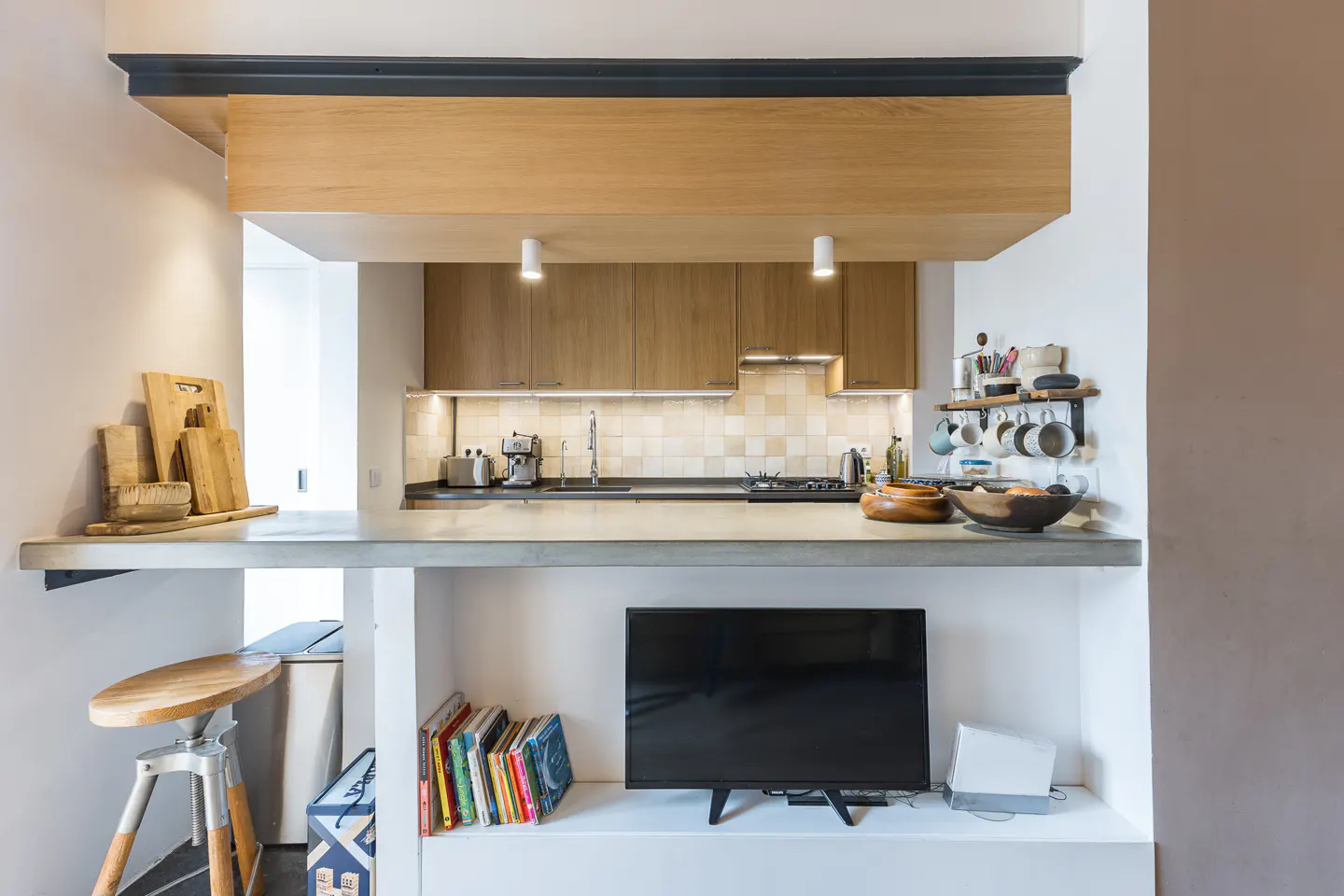 Modern kitchen with wood cabinets, concrete breakfast bar, TV on a lower shelf, and a wooden stool.