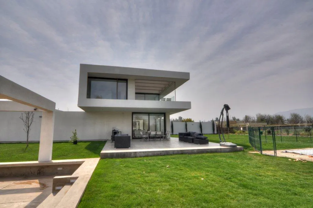 Modern two-story white house with a patio, outdoor furniture, and green lawn under a cloudy sky.