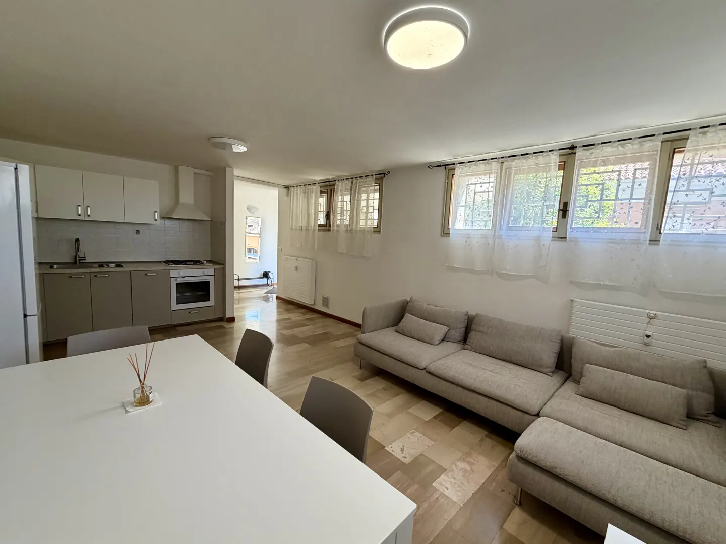 Open-concept living space with a kitchen, dining table, and a gray sectional sofa near windows with sheer curtains.