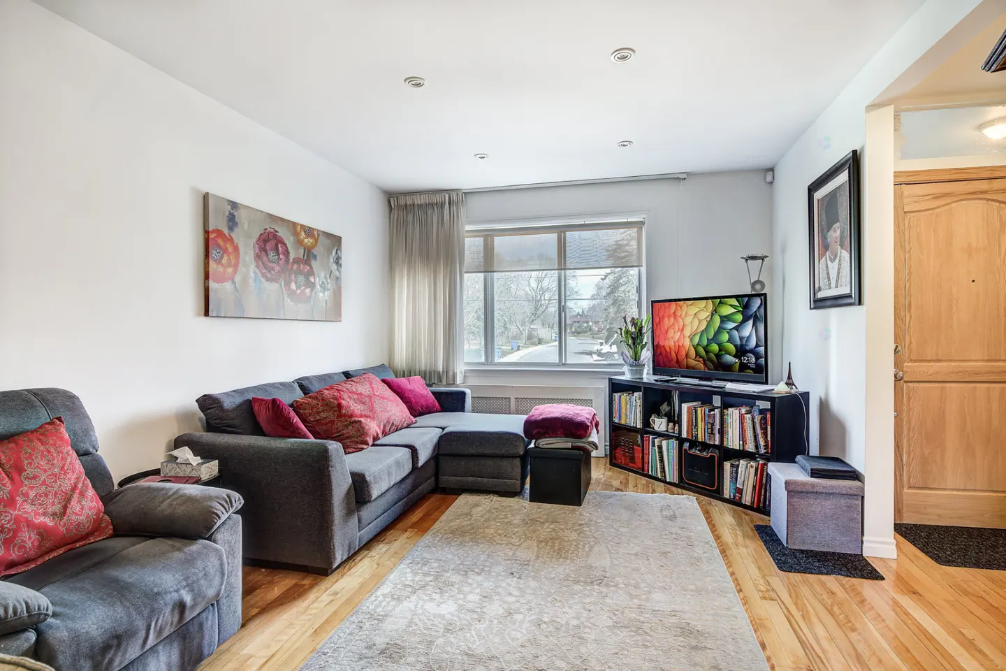 Living room with gray sectional sofa, red pillows, and a large window. A TV sits on a bookshelf. Hardwood floors and a light gray rug complete the space.