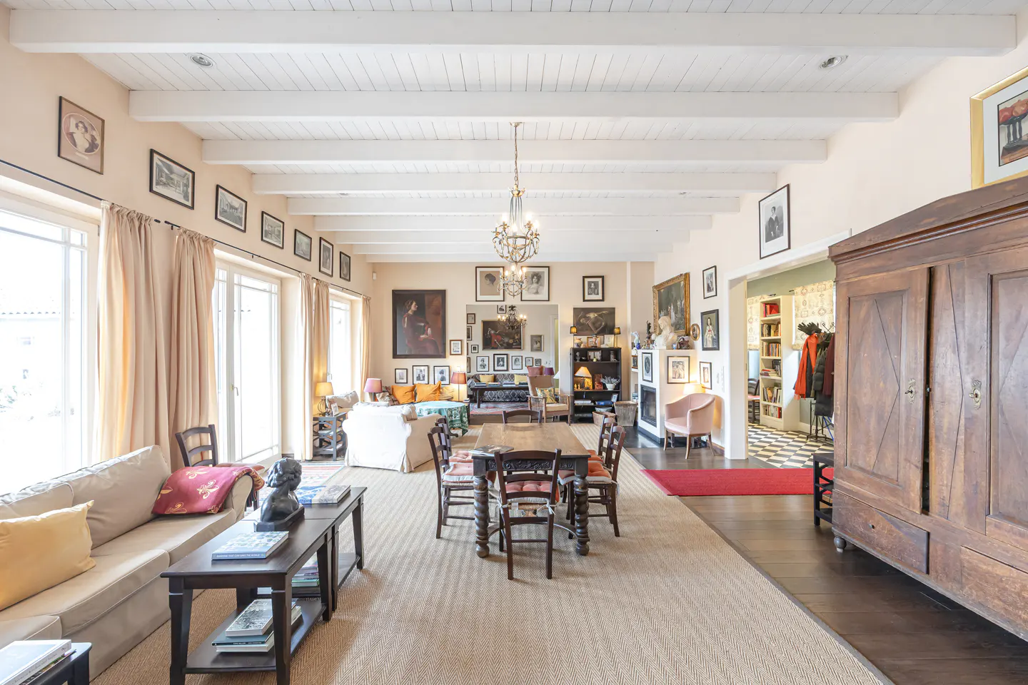 Spacious living room with beige walls, white beamed ceiling, wood floors, and a large area rug. A dining table and sofa are visible.