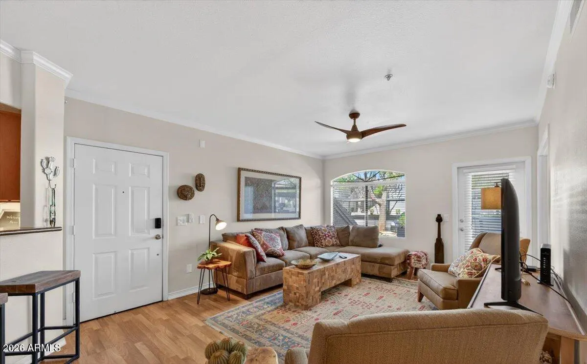 A bright living room with a brown sectional sofa, armchairs, and a patterned rug on a wood floor. A ceiling fan is visible.