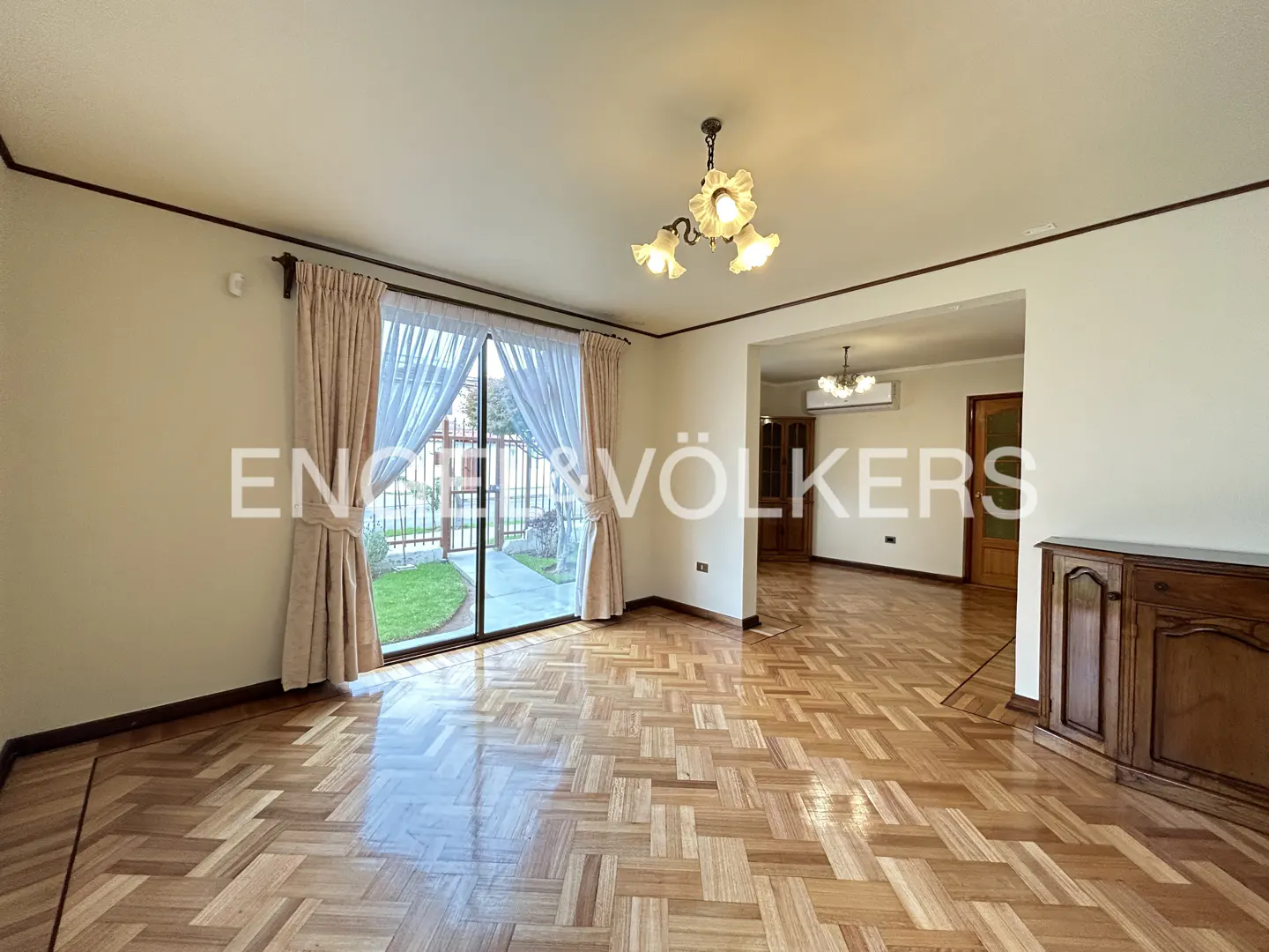 A bright living room with parquet floors, beige walls, and a glass door leading to a garden. A wooden cabinet stands against the wall.
