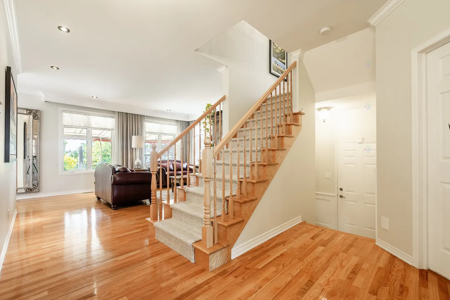 Bright foyer with hardwood floors, a carpeted staircase with wooden banister, and a glimpse of a living room with leather furniture.