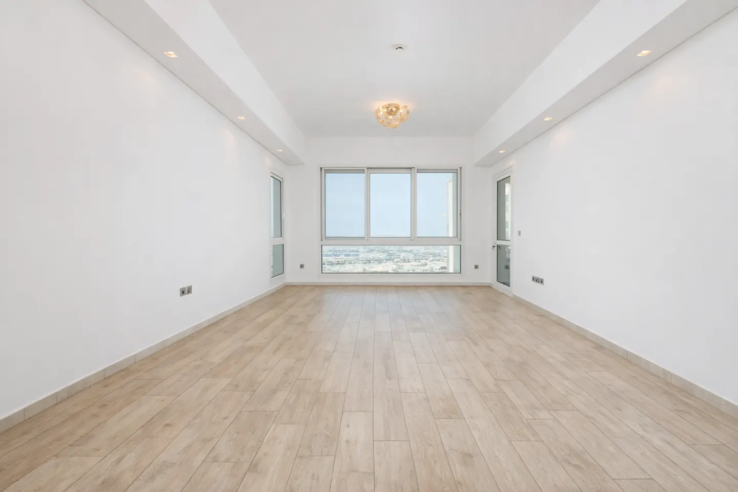 Bright, empty room with light wood floors, white walls, and a large window with a city view. A chandelier hangs from the ceiling.