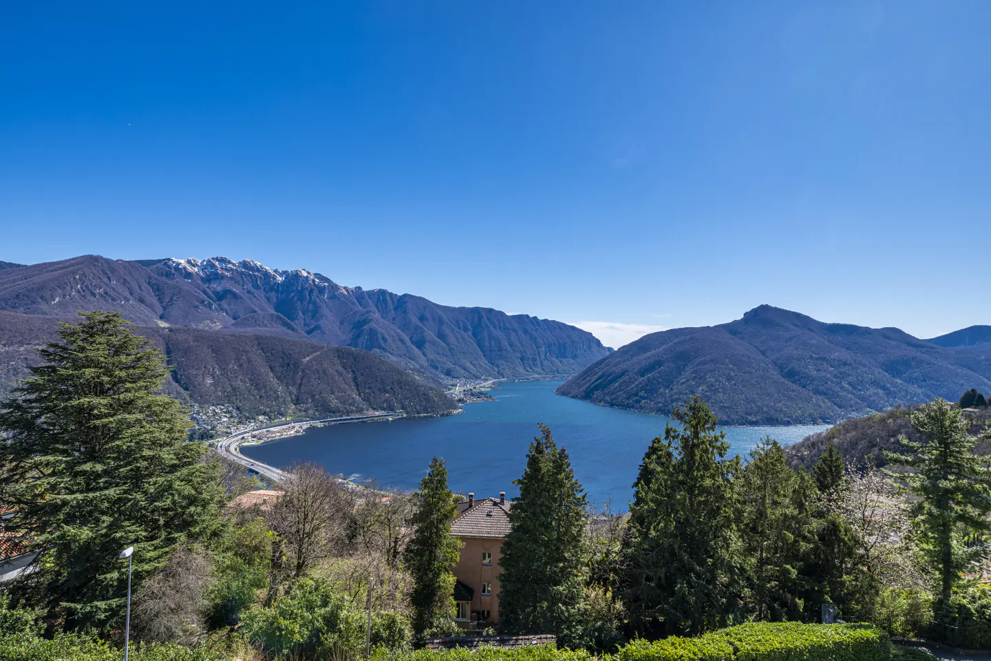 Scenic view of Lake Lugano, Switzerland, with blue water, green trees, and snow-capped mountains under a clear blue sky.