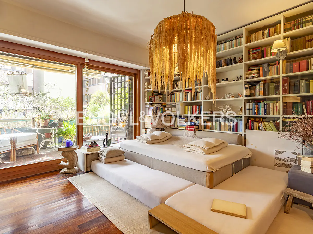 Living room with a large bookshelf, a tiered platform bed, and a balcony with plants. A unique chandelier hangs above.