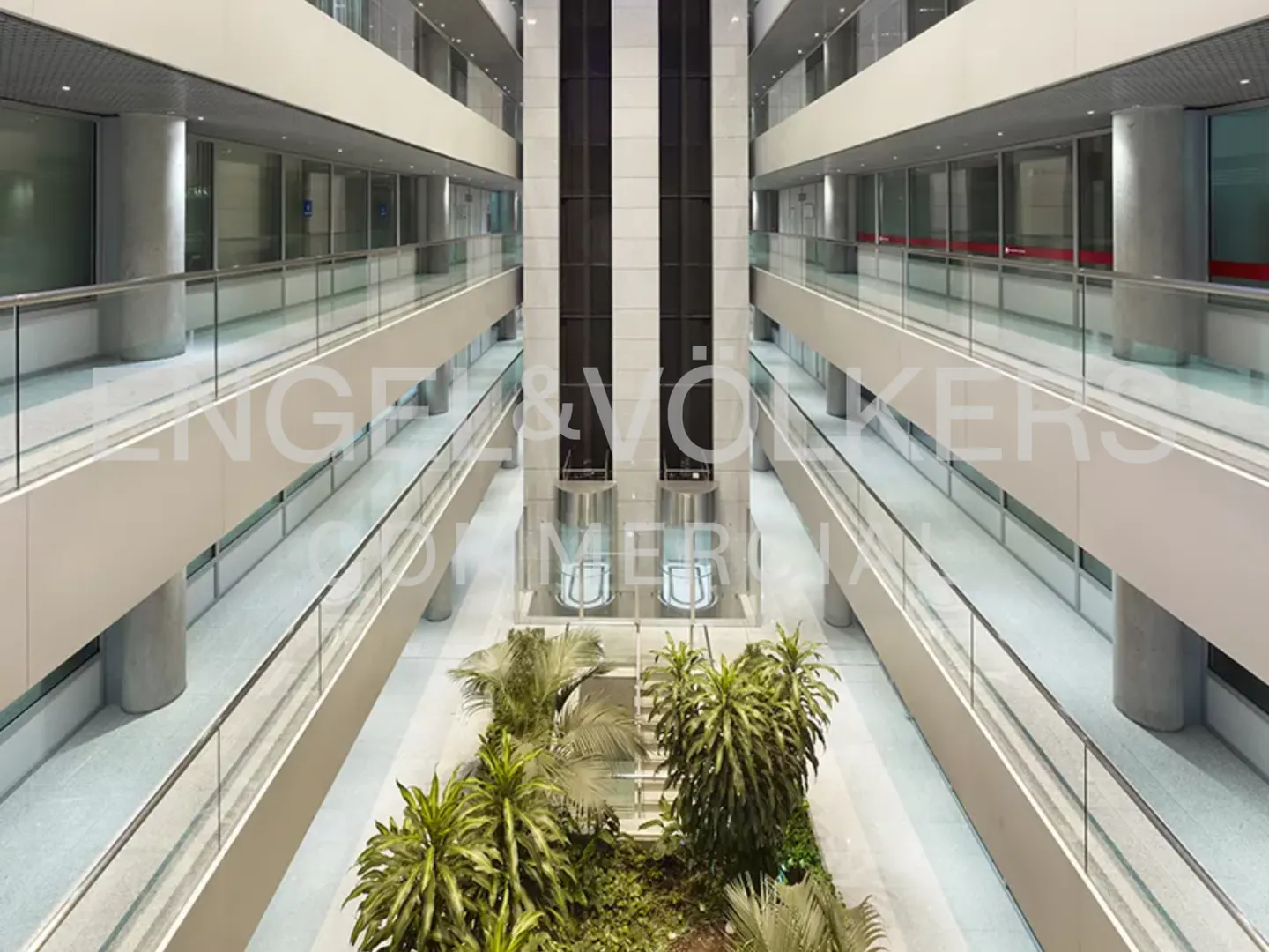 Interior view of a modern office building with multiple floors, glass railings, elevators, and a green plant display on the ground floor.