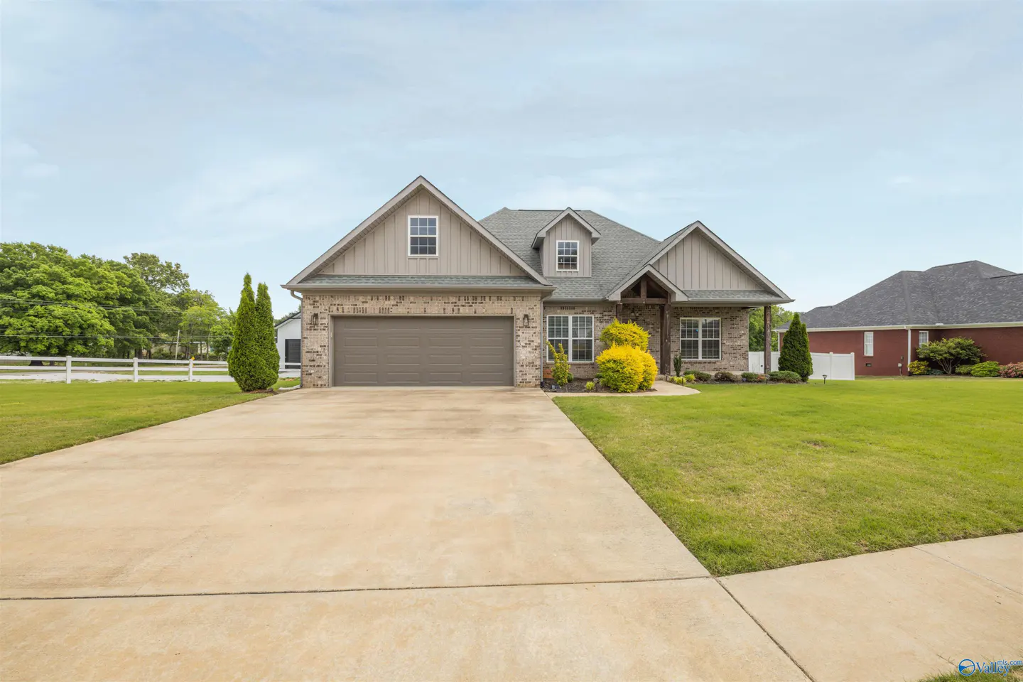 Tan brick house with a gray roof, a two-car garage, and a long driveway on a green lawn under a blue sky.