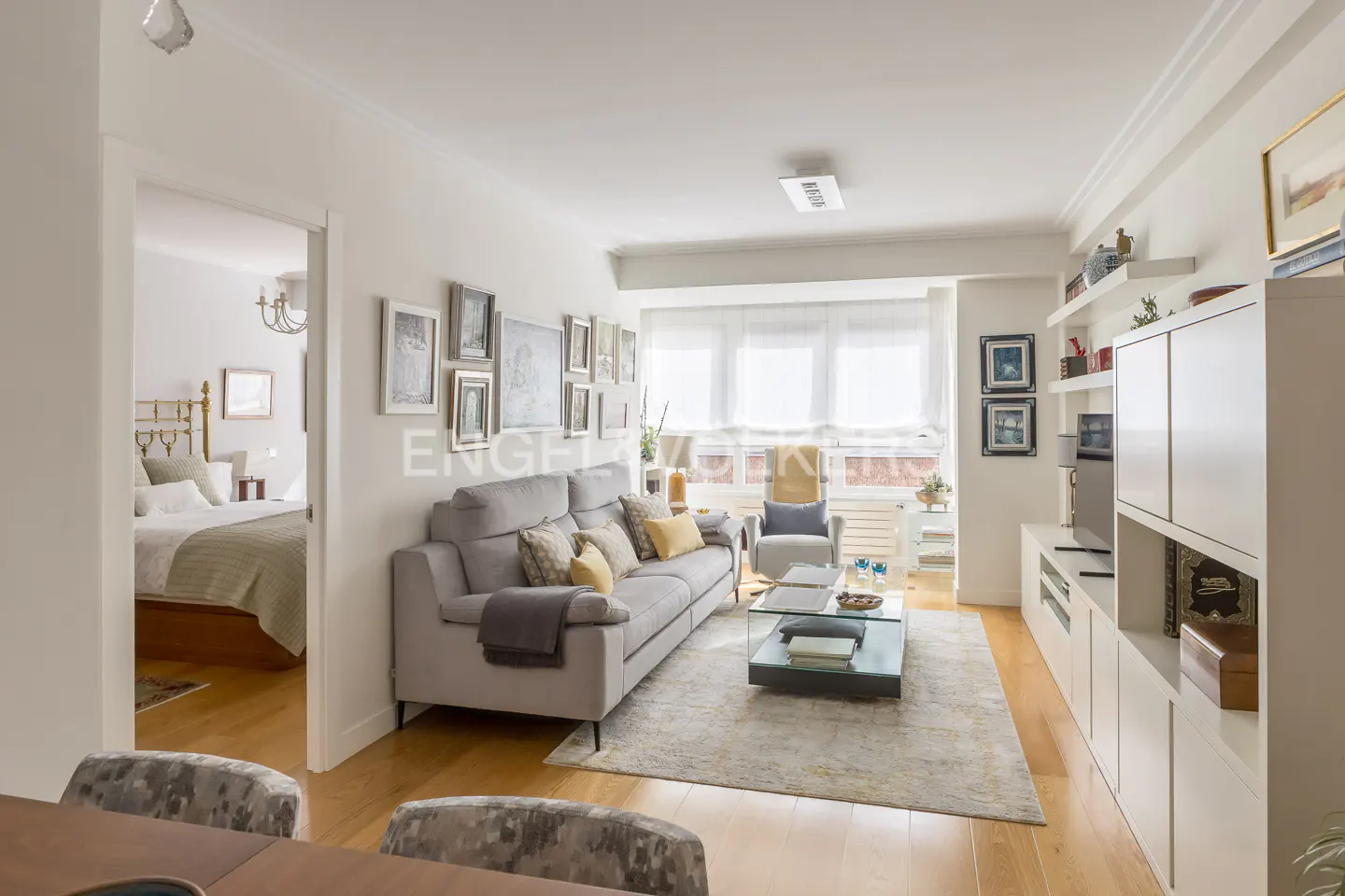 Bright living room with a grey sofa, glass coffee table, and white shelving, with a view into a bedroom with a brass bed.