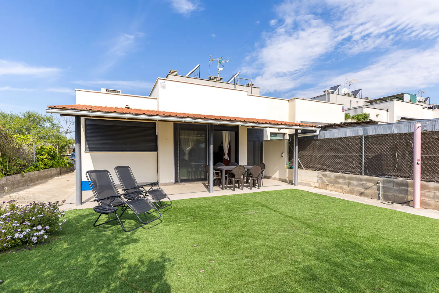 Backyard view of a modern white house with a red roof, green lawn, lounge chairs, and outdoor dining set.
