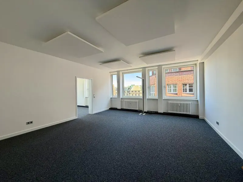 Bright, empty office space with white walls, gray carpet, and large windows showing a brick building. Rectangular soundproofing panels hang from the ceiling.