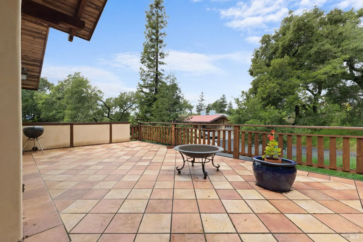 Tiled patio with a grill, fire pit, and potted plant. A wooden fence surrounds the patio with trees in the background.