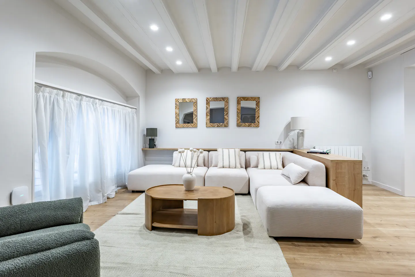 Bright living room with white walls, wood floors, and beamed ceiling. A large white sectional sofa faces a round wood coffee table on a light rug. Three square mirrors hang above the sofa. Curtains cover a window.