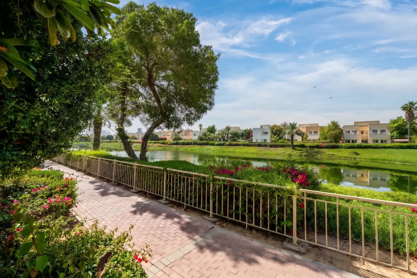 A paved walkway runs alongside a calm lake, bordered by a white fence, lush greenery, and pink flowers under a blue sky.