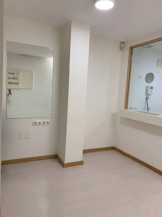 Bright, empty room with white walls, light wood floors, and a large mirror. A window with a view into another room is on the right.