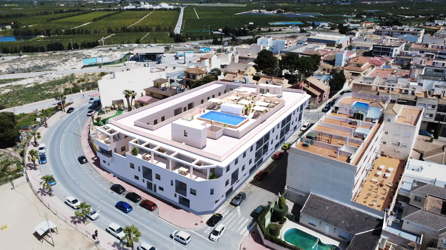 Aerial view of a modern white building with a rooftop pool and lounge area, surrounded by a coastal town and green fields.