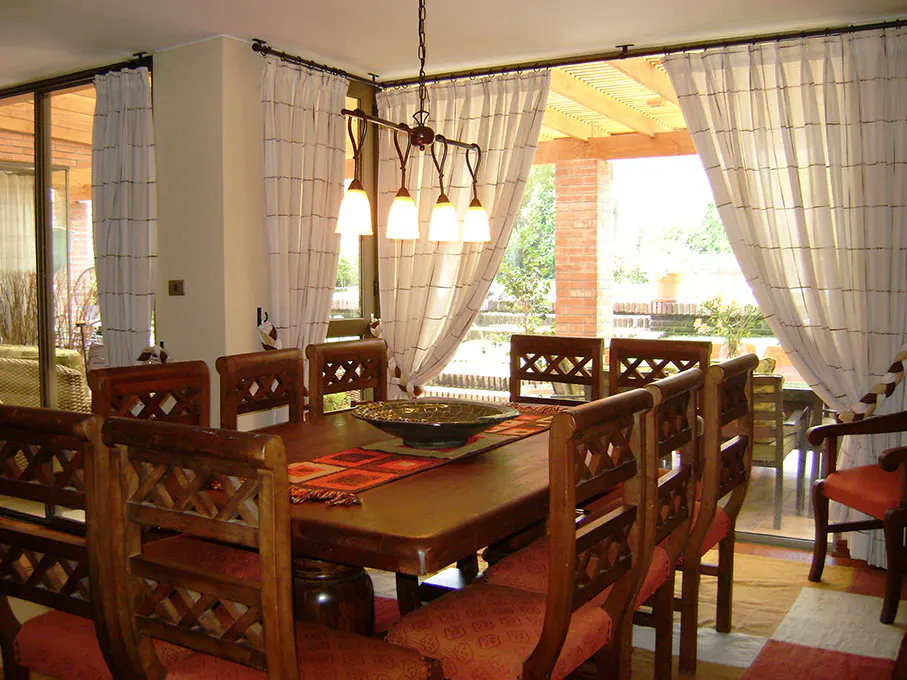 Dining room with a long wooden table, chairs with red cushions, and a hanging light fixture. White curtains frame a view of an outdoor patio.