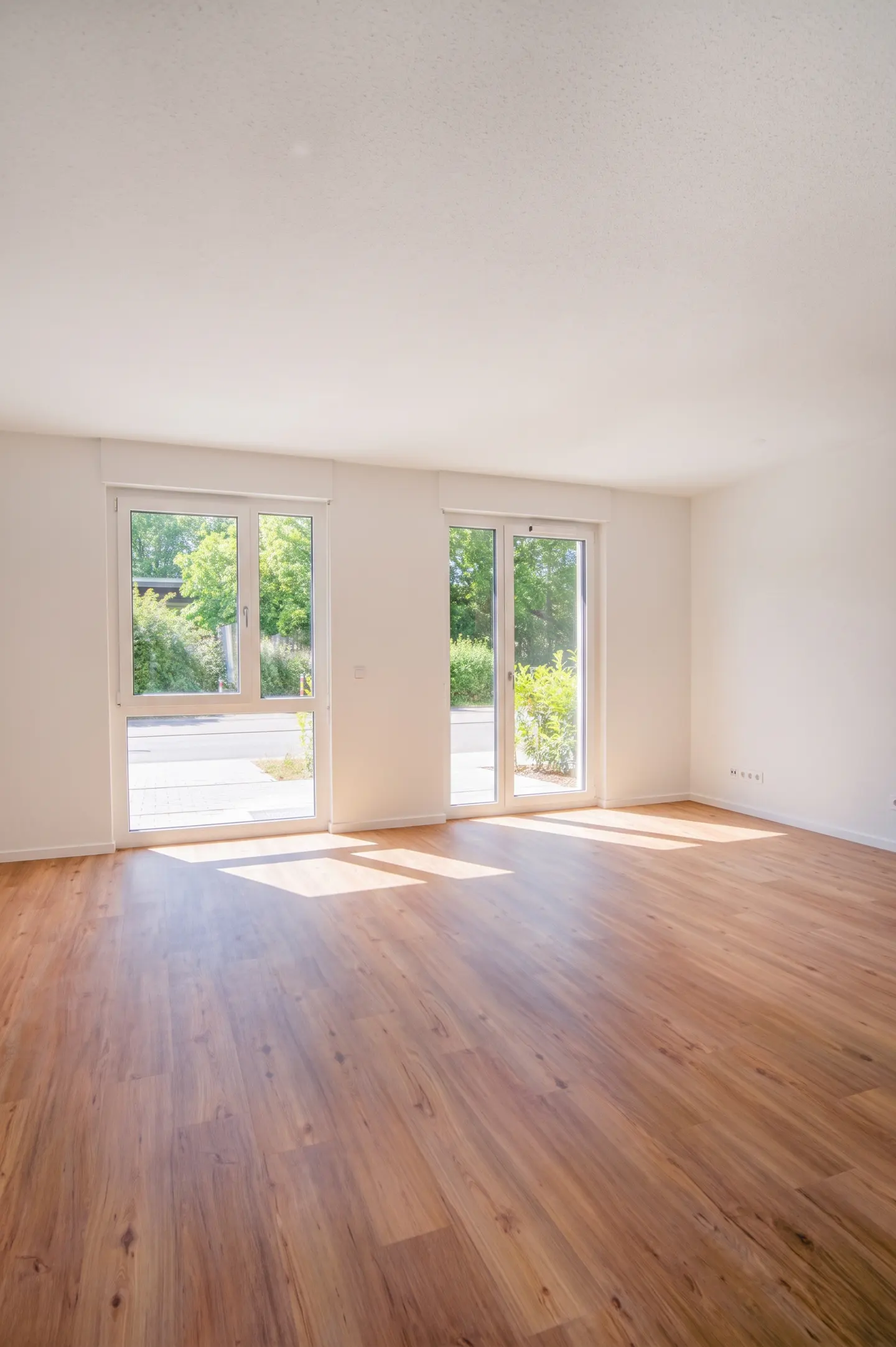 Bright, empty room with wood floors, white walls, and two large windows showing green trees outside.