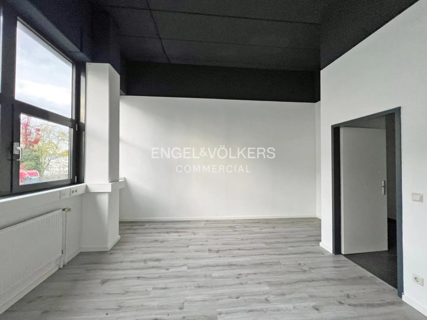 Empty commercial space with gray wood floors, white walls, black ceiling, and Engel & Völkers logo. A window and doorway are visible.