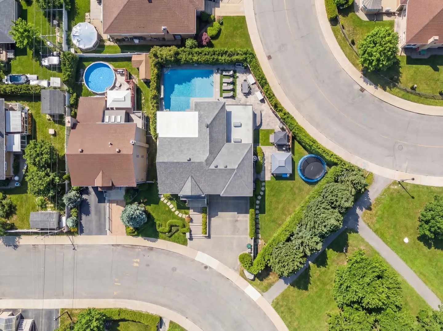 Aerial view of a grey roofed house with a pool, trampoline, and green lawn in a suburban neighborhood.