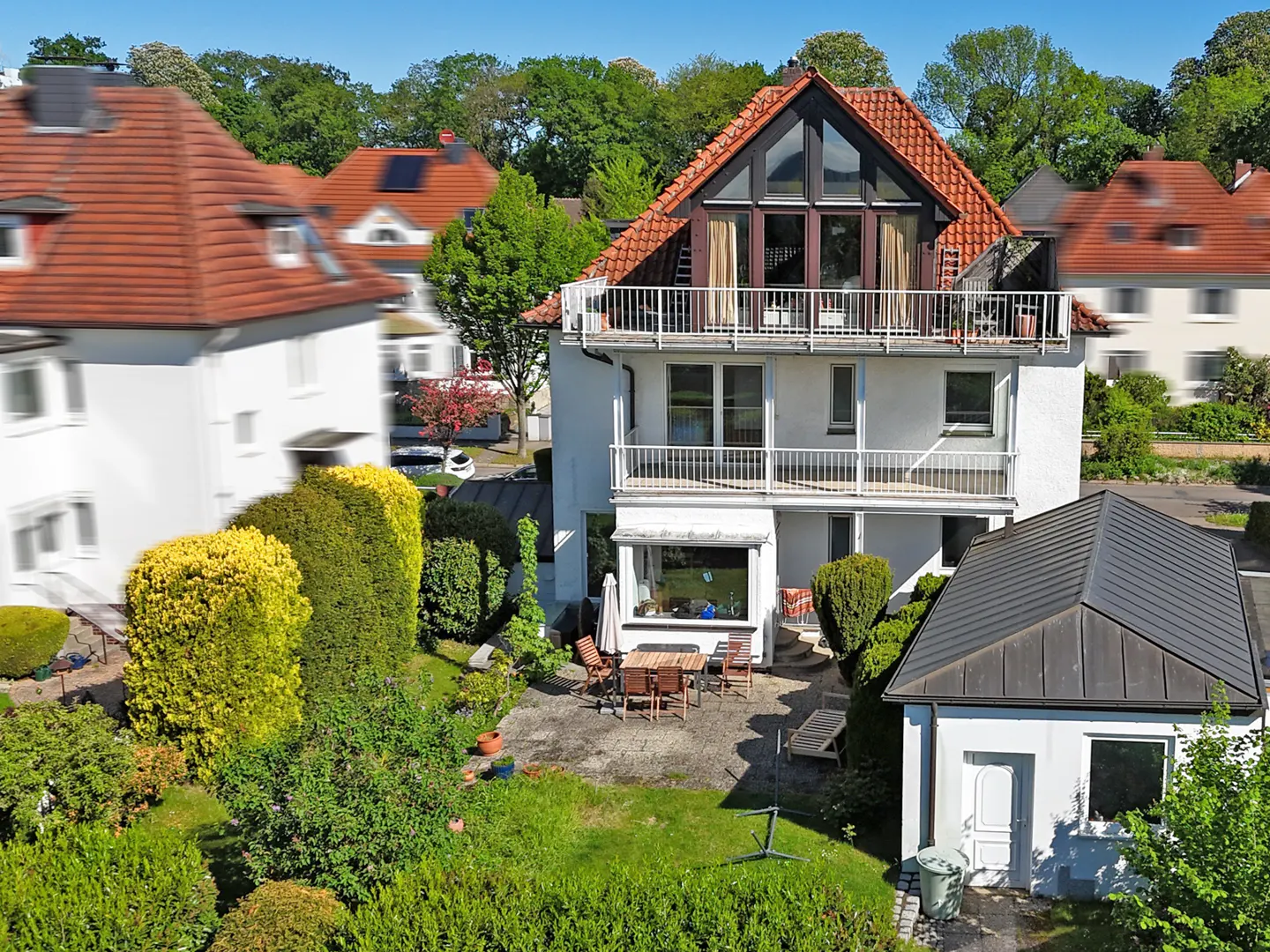 A three-story white house with a red tile roof and two balconies, surrounded by green trees and bushes. A patio with a table and chairs is visible.