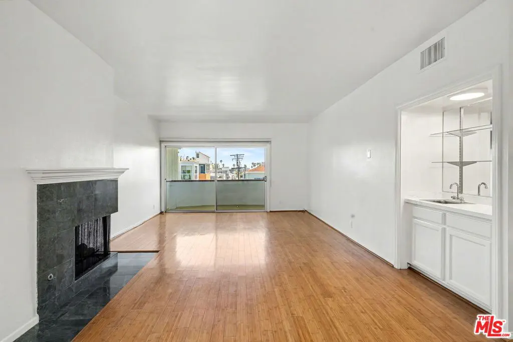 Bright living room with hardwood floors, white walls, and a black marble fireplace. Sliding glass doors lead to a balcony. A wet bar is built into the wall.