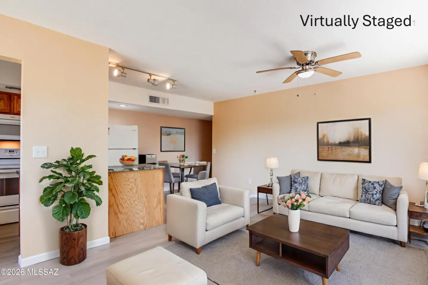 A staged living room with beige walls, a white sofa, a chair, and a dark wood coffee table. A kitchen is visible in the background.