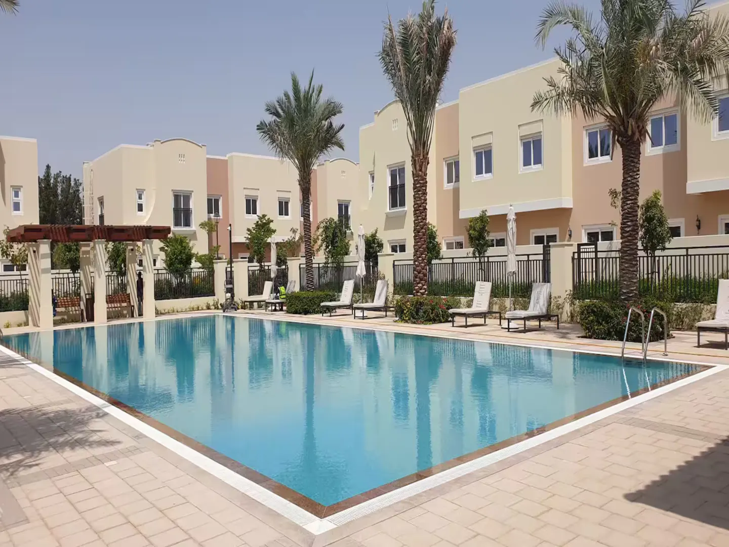 A rectangular swimming pool with clear blue water, surrounded by beige-colored townhouses and palm trees.