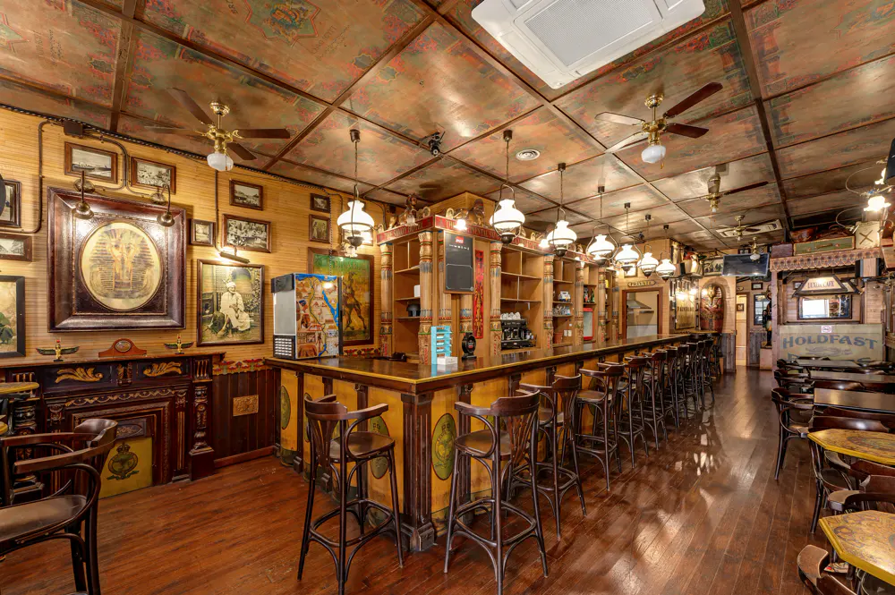 A rustic pub interior with a long wooden bar, high stools, ornate ceiling panels, and walls decorated with framed art.