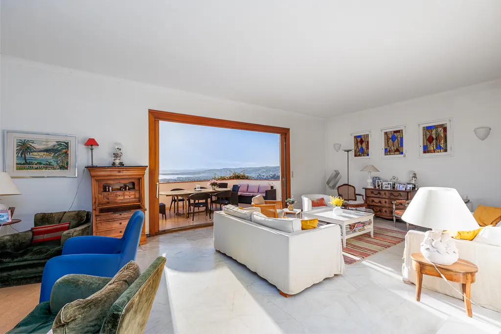 Bright living room with white walls, marble floors, and a large sliding door to a patio with ocean views. Sofas, chairs, and wooden furniture fill the space.