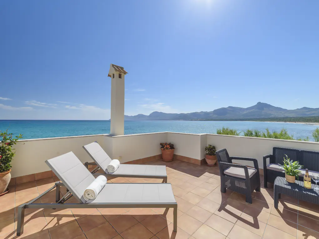 Rooftop terrace with ocean view. Two lounge chairs, table and chairs, and potted plants. Blue sky and water.