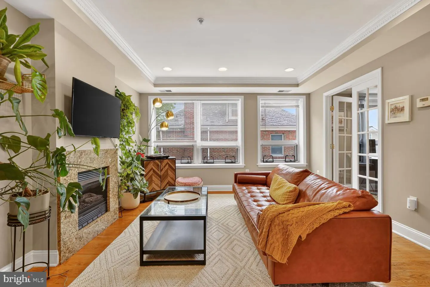 A living room with a brown leather sofa, a glass coffee table, and a fireplace with a TV above it. There are plants around the fireplace.