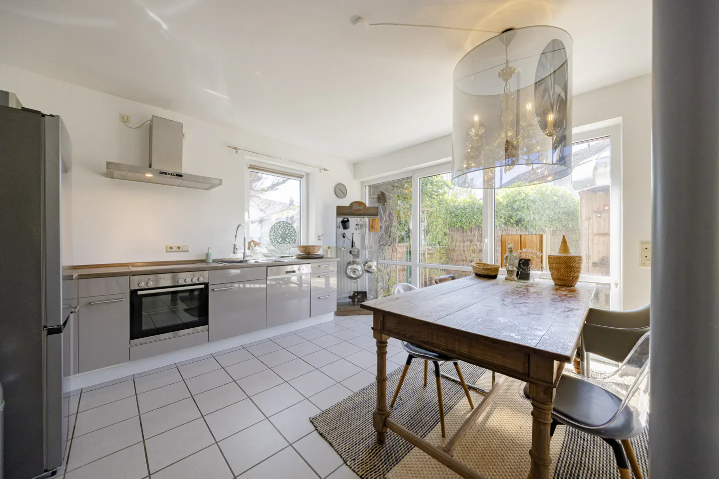 Bright kitchen with gray cabinets, white tile floor, and a wooden table with clear chairs under a chandelier.