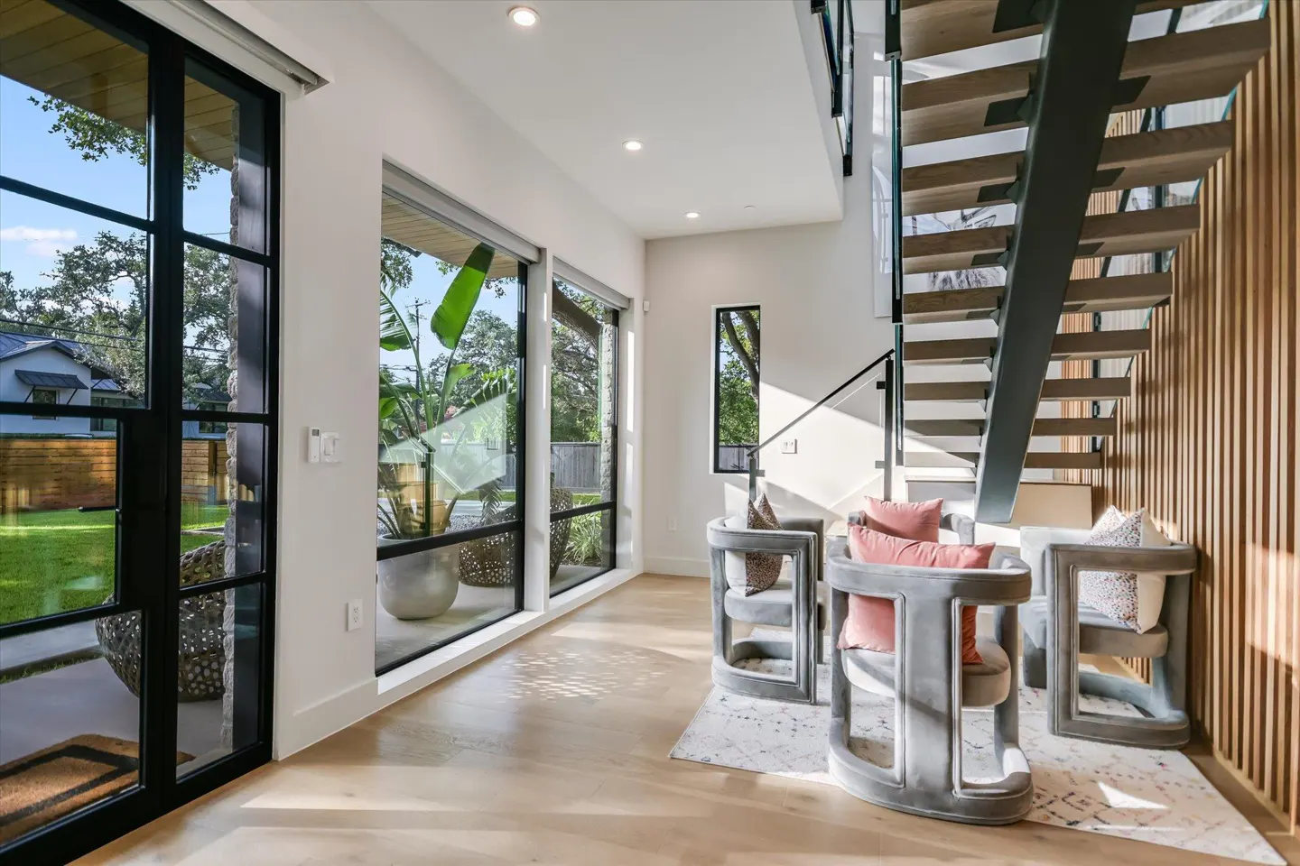 Bright, modern foyer with black-framed windows, light wood floors, and three gray chairs with pink pillows under a floating staircase.