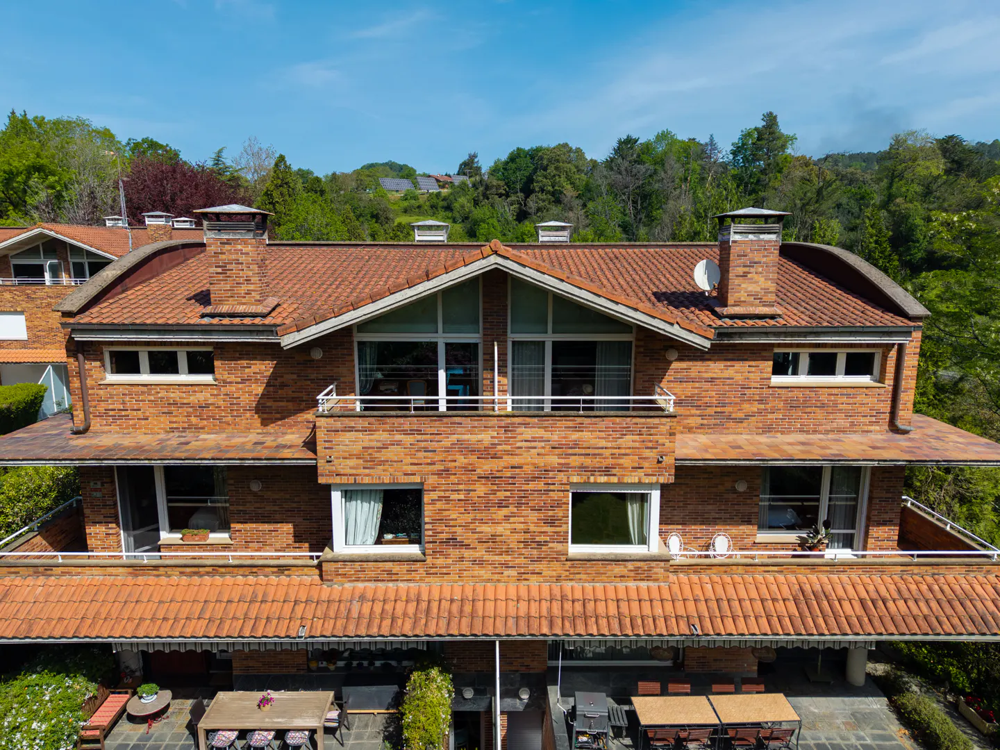 Aerial view of a two-story brick house with a red tile roof, chimneys, and outdoor seating areas. Green trees and blue sky in the background.
