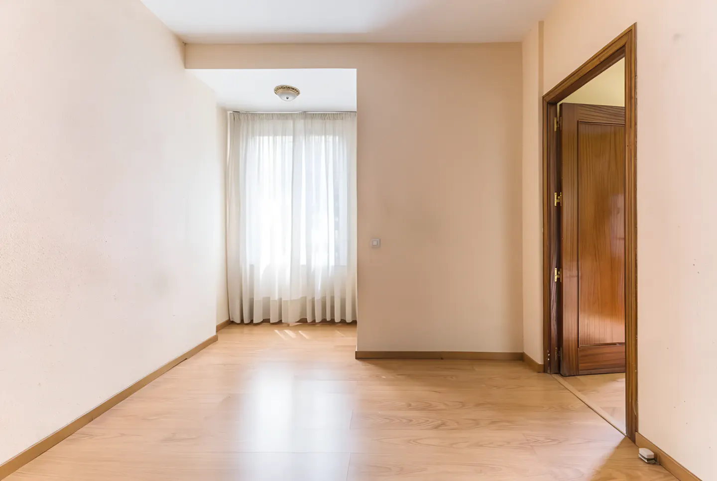 Empty room with light wood floors, beige walls, and a window with sheer white curtains. Ajar wood door on the right.