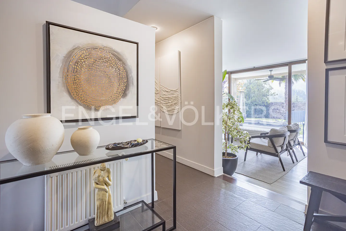 A modern home's entryway with a glass console table, white vases, and abstract art. A view to an outdoor patio with seating.