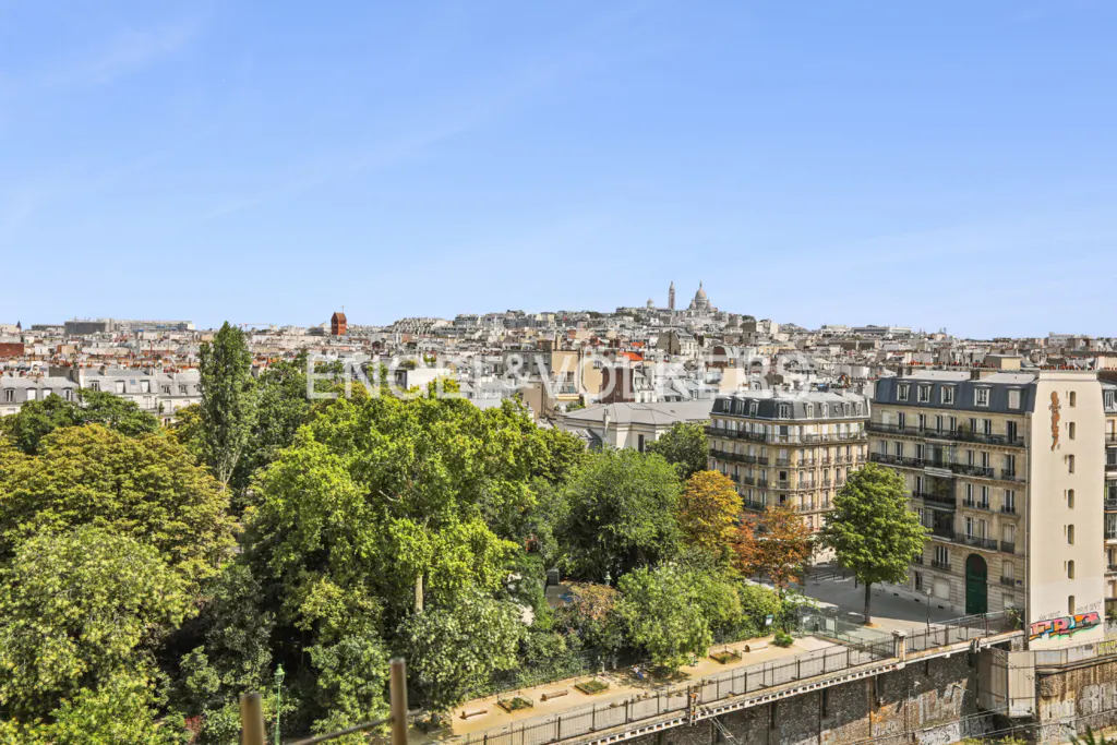 Paris cityscape view with green trees, buildings, and the Sacré-Cœur Basilica in the distance under a clear blue sky.