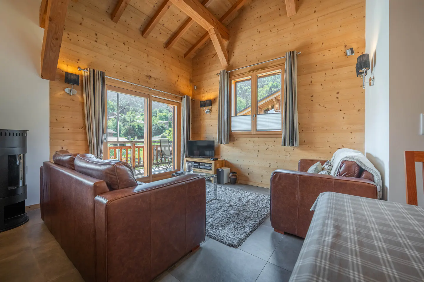 Cozy living room with wood walls, beams, and leather sofas. A fireplace, TV, and balcony with mountain views add to the chalet's charm.